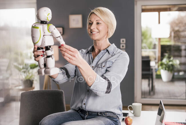 Улыбающаяся деловая женщина, держащая в руках модель робота, сидя за столом в домашнем офисе — стоковое фото