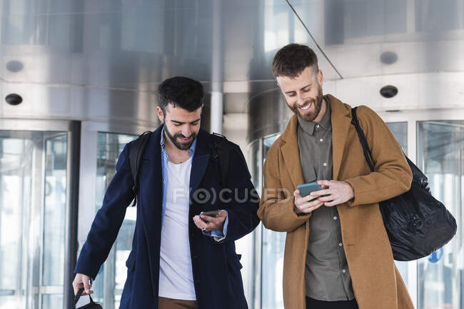 Parceiros de negócios usando telefones celulares enquanto estão de pé com saco contra edifício — Fotografia de Stock