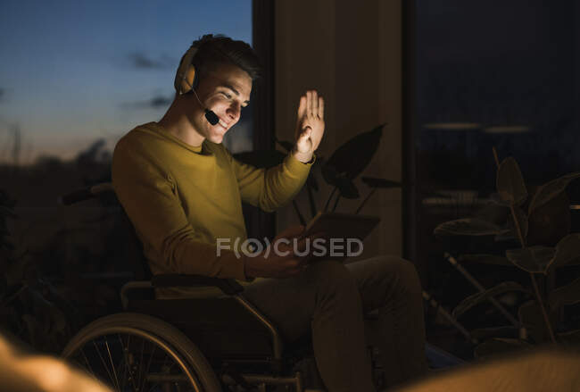 Smiling young man in wheelchair gesturing while making video call through digital tablet sitting on wheelchair in living room — Stock Photo