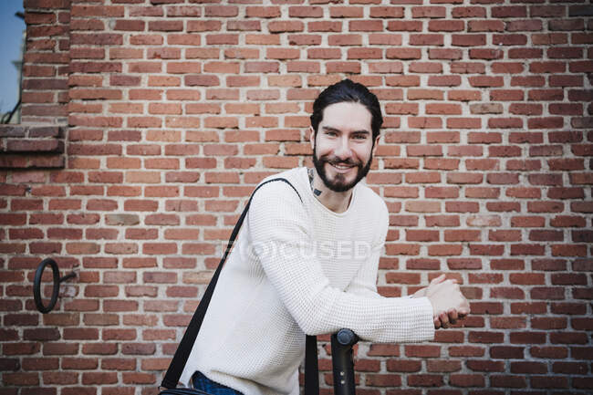 Sonriente hombre con las manos apretadas apoyándose en la vespa de empuje por la pared de ladrillo - foto de stock