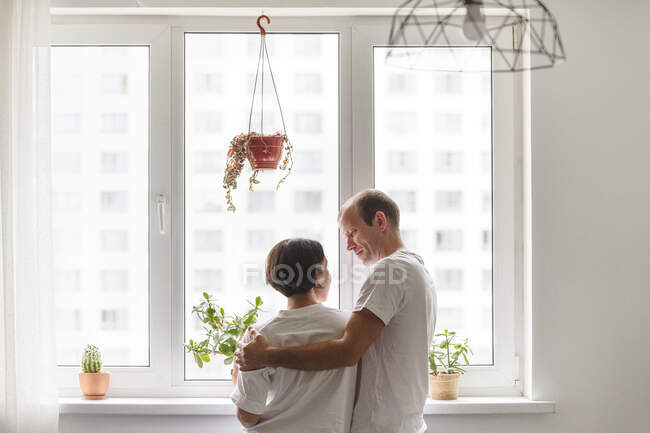 Man and woman looking at each other while embracing near window at home — Stock Photo
