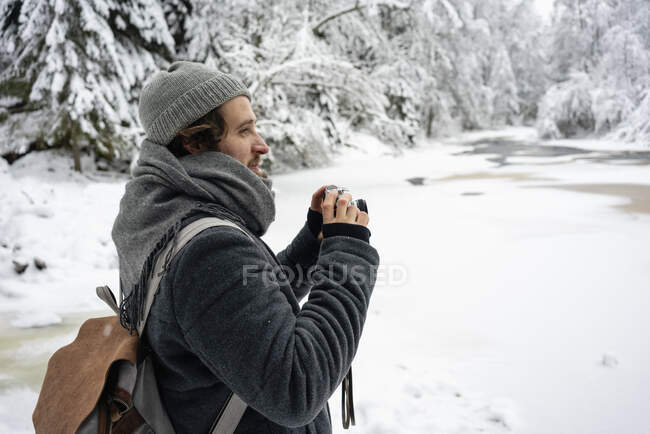 Joven con mochila y cámara mirando hacia otro lado mientras está parado en el bosque - foto de stock