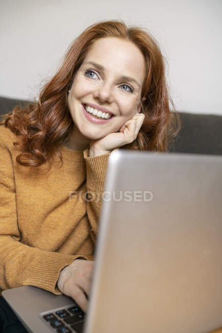 Hermosa pelirroja sonriente pensando mientras mira hacia otro lado en casa - foto de stock