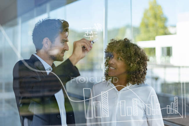 Männerprofi diskutiert mit Kollegin über Grafik auf Glas im modernen Büro — Stockfoto