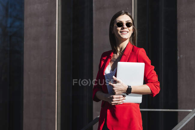 Smiling businesswoman with sunglasses holding laptop while standing against brown structure — Stock Photo
