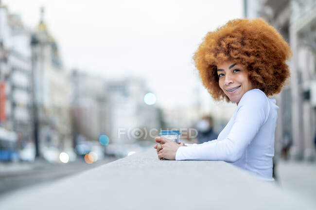 Улыбающаяся кудрявая женщина, стоящая с одноразовой чашкой на улице — стоковое фото
