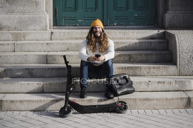 Laughing male hipster with mobile phone sitting on steps during sunny day — long  hair, scooter - Stock Photo | #490890004