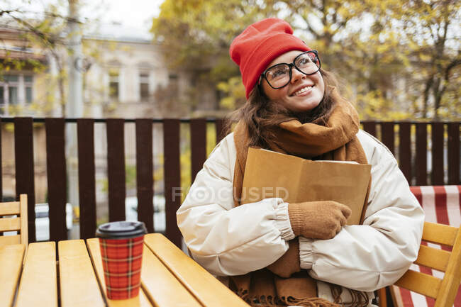 Femme réfléchie souriant tout en embrassant livre assis au café trottoir — Photo de stock