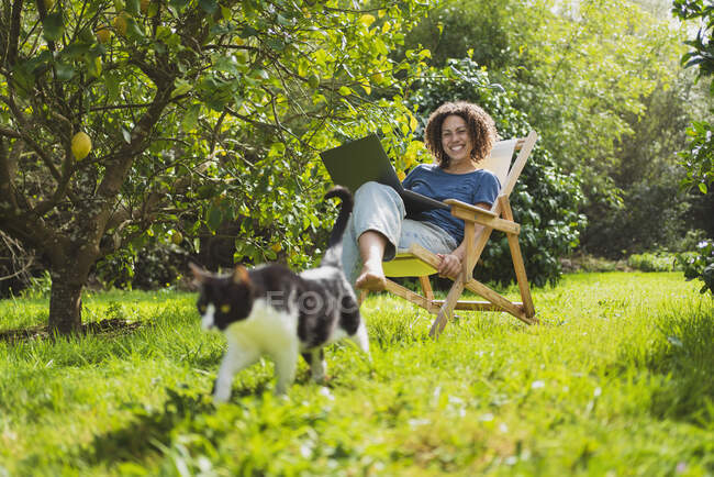 Happy woman with laptop looking at cat while sitting on deck chair by lemon tree — Stock Photo