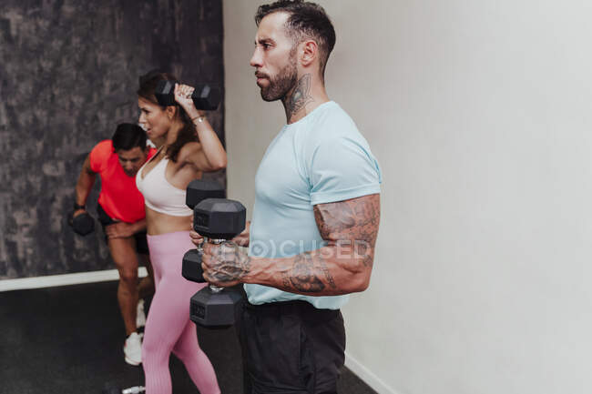 Sportsman holding dumbbell while standing with friends exercising in background at gym — Stock Photo