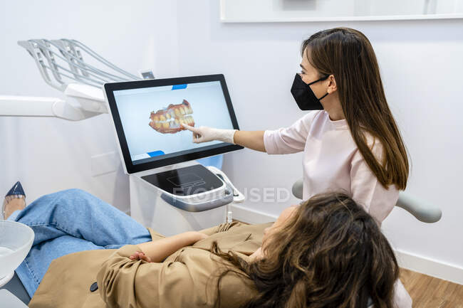Dentista femenina explicando en pantalla de computadora a paciente en clínica durante pandemia - foto de stock