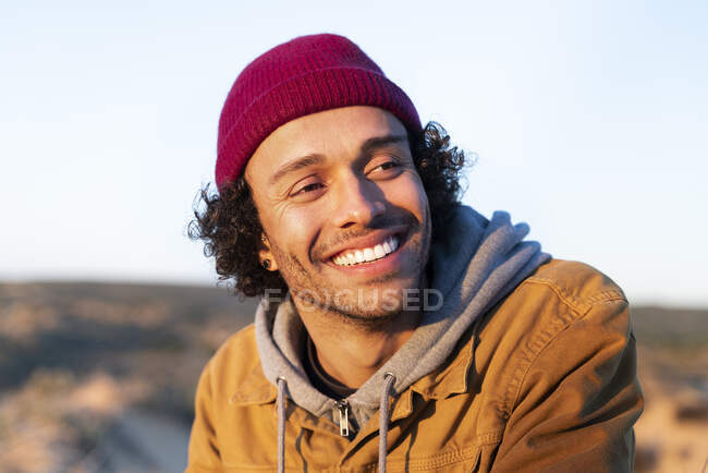 Усміхнений чоловік у в'язаному капелюсі дивиться вбік, сидячи на відкритому повітрі — стокове фото