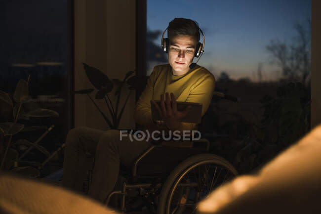 Jeune homme en fauteuil roulant portant un casque sans fil tout en utilisant une tablette numérique assis sur fauteuil roulant dans le salon — Photo de stock