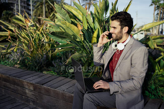 Uomo d'affari sorridente con computer portatile che parla sul telefono cellulare mentre si siede sul muro di contenimento — Foto stock