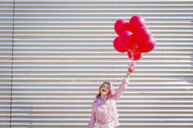 Mujer feliz con los ojos cerrados sosteniendo racimo en globos delante de la pared de metal - foto de stock