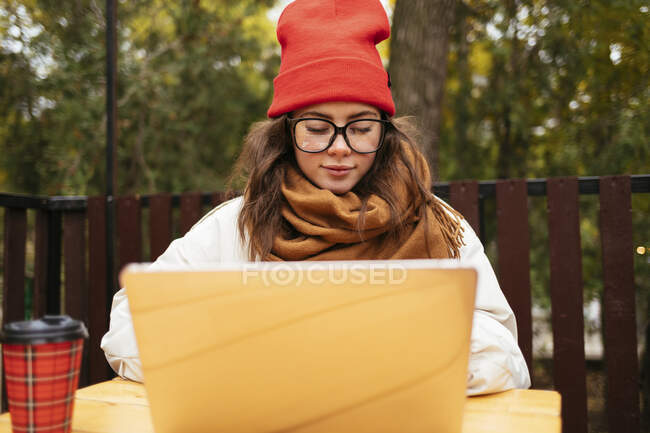 Женщина в вязаной шляпе и шарфе с помощью ноутбука, сидя в кафе на тротуаре — стоковое фото