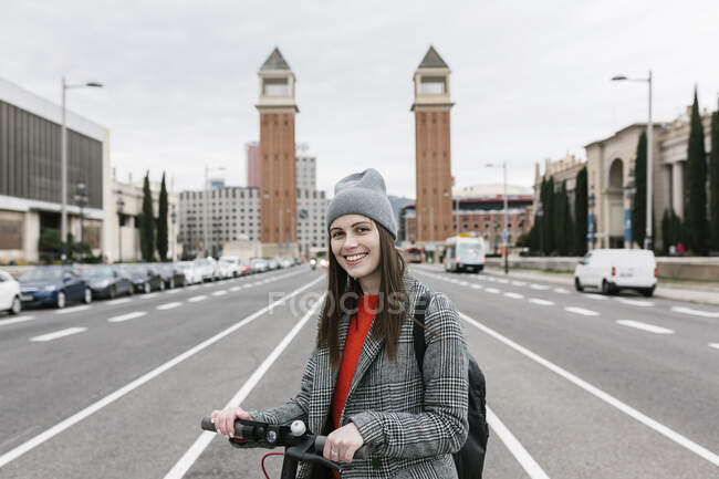 Donna sorridente con scooter elettrico spinta in piedi sulla strada contro Torres Venecianes in città — Foto stock