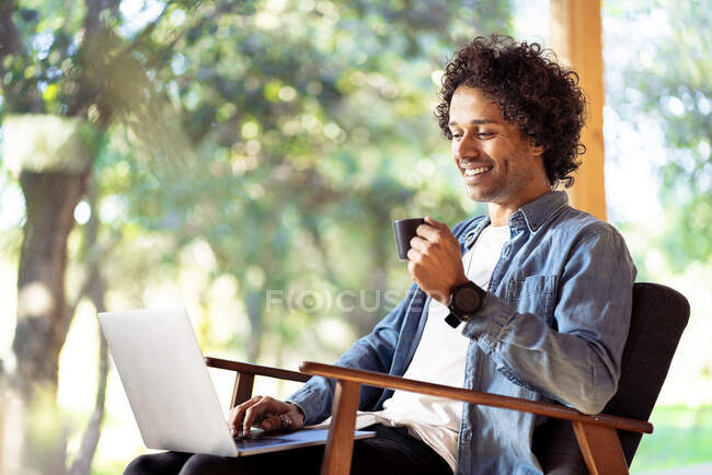 Посмішка чоловіка з чашкою кави з ноутбуком під час сидіння на кріслі на подвір'ї. — стокове фото