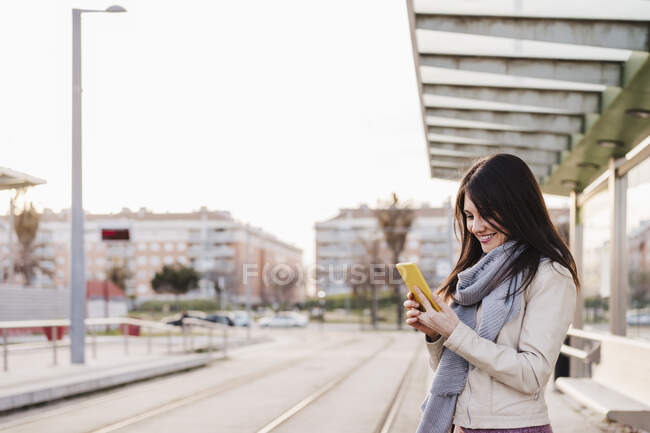 Усміхнена жінка у шарфі за допомогою смартфона стоїть на залізничній станції. — стокове фото