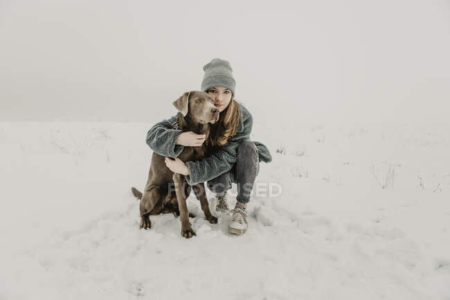 Portrait of teenage girl crouching in snow and embracing Labrador Retriever — Stock Photo