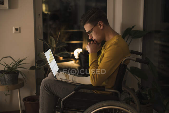 Homme concentré en fauteuil roulant avec ordinateur portable travaillant tard tout en étant assis en fauteuil roulant dans le salon — Photo de stock