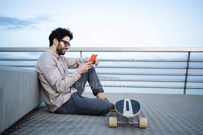 Hombre de negocios sonriente usando el teléfono móvil por longboard mientras está sentado en el muelle apoyado contra el banco - foto de stock