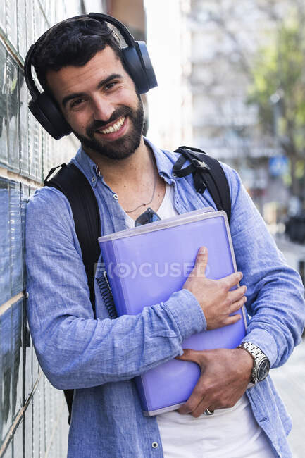 Feliz hombre de negocios con documentos escuchando música mientras se apoya en la pared - foto de stock