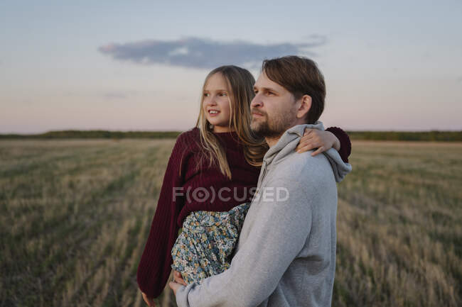 Padre llevando a su hija de pie en el campo viendo la puesta de sol - foto de stock