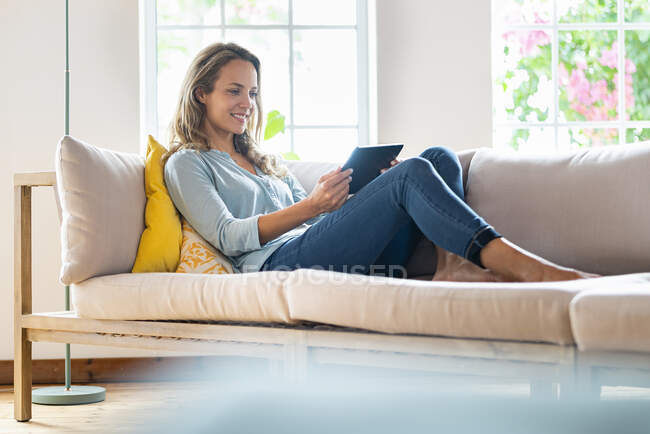 Femme souriante dans les occasionnels regardant tablette numérique tout en étant assis sur le canapé — Photo de stock