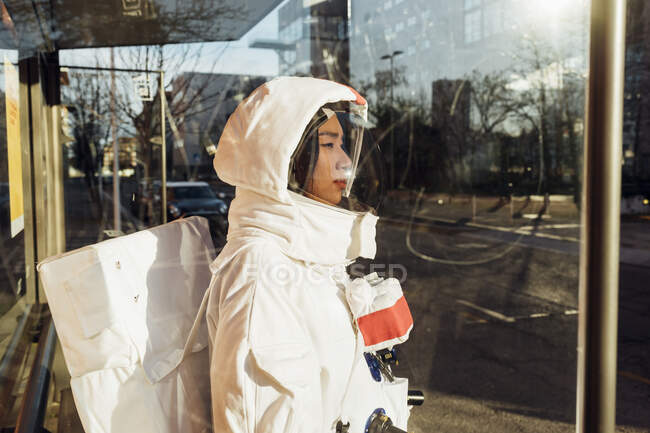 Astronauta donna in tuta spaziale in piedi alla fermata dell'autobus durante la giornata di sole visto attraverso il vetro — Foto stock
