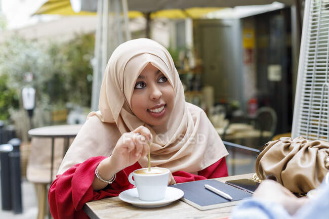 Sorridente giovane donna mescolando caffè mentre seduto al caffè marciapiede — Foto stock