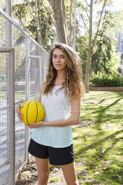 Young beautiful woman holding basketball while standing by fence — Stock Photo