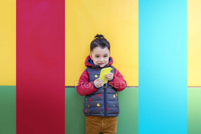 Sonriente niño con el teléfono móvil de pie delante de la pared colorida - foto de stock