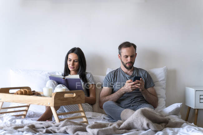 Woman reading book sitting with man using smart phone while having breakfast on bed at home — Stock Photo