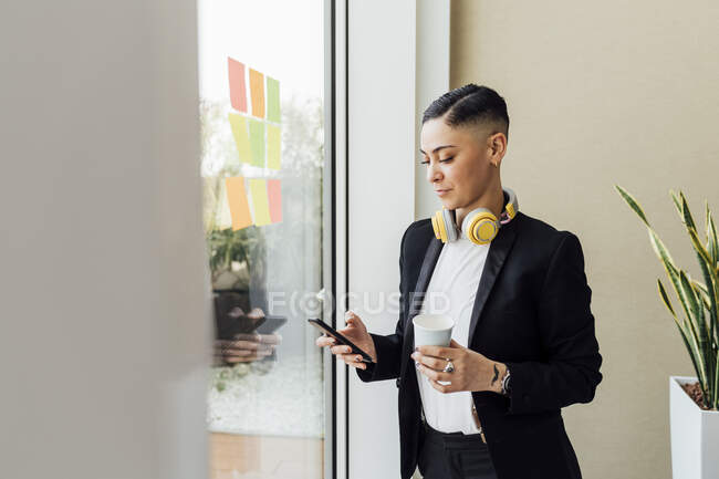Жінка-підприємець тримає одноразову чашку кави під час використання смартфона в офісі — стокове фото