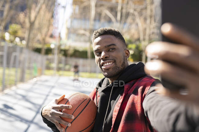 Молодой человек проводит баскетбол, делая селфи на мобильном телефоне в солнечный день — стоковое фото
