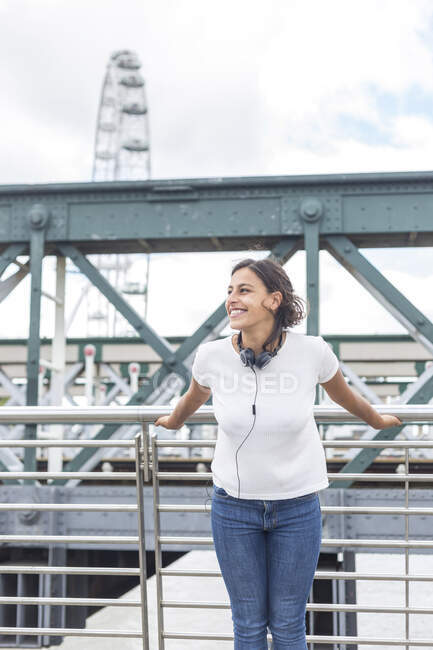 Femme heureuse avec écouteurs debout devant la rampe sur le pont, Londres, Angleterre, Royaume-Uni — Photo de stock