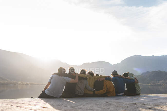 Amici maschi e femmine seduti sul lungomare con le braccia intorno durante la giornata di sole — Foto stock