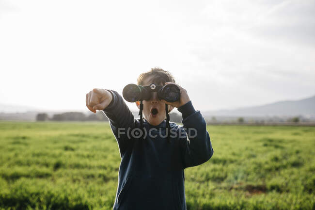 Surprised boy pointing while looking through binoculars in field during sunny day — Stock Photo