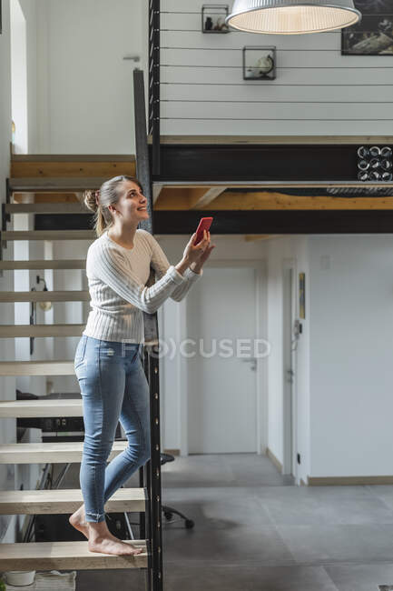 Donna sorridente che controlla le funzioni domestiche con lo smartphone mentre è in piedi su gradini a casa — Foto stock