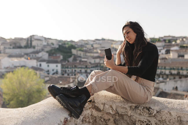Turista femenina tomando selfie a través del teléfono móvil mientras está sentada en la pared de contención - foto de stock