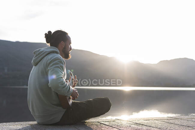 Uomo giocare ukulele mentre seduto sul lungomare vicino al lago — Foto stock