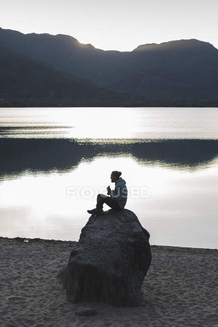 Uomo seduto sulla roccia al lago durante il tramonto — Foto stock