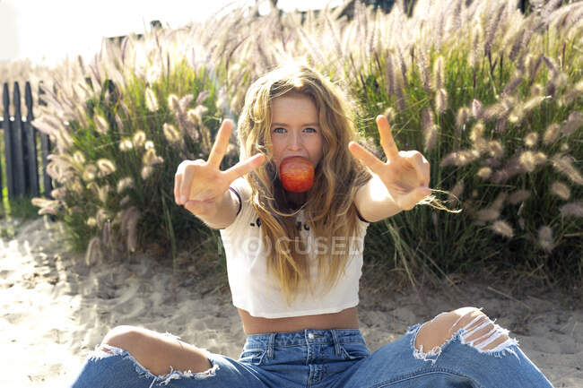 Frau isst Apfel und zeigt Friedenszeichen, während sie an sonnigen Tagen auf Sand sitzt — Stockfoto