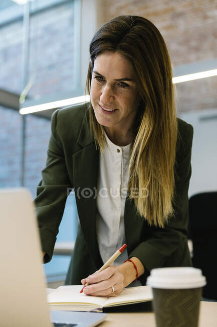 Взрослая деловая женщина, пишущая заметки в дневнике во время работы на ноутбуке в офисе — стоковое фото