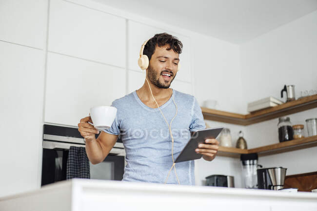 Young man holding coffee cup and tablet while listening music through headphones in kitchen — Stock Photo