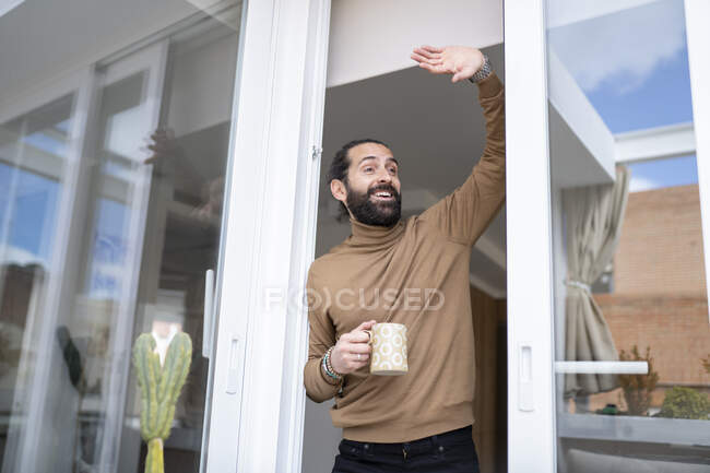 Feliz hombre saludando mientras está de pie por la puerta corredera en casa - foto de stock