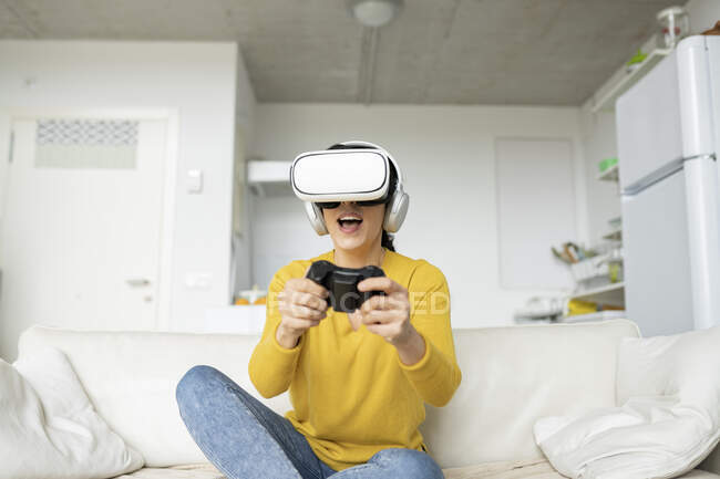 Donna che gioca indossando cuffie realtà virtuale mentre seduto sul divano a casa — Foto stock