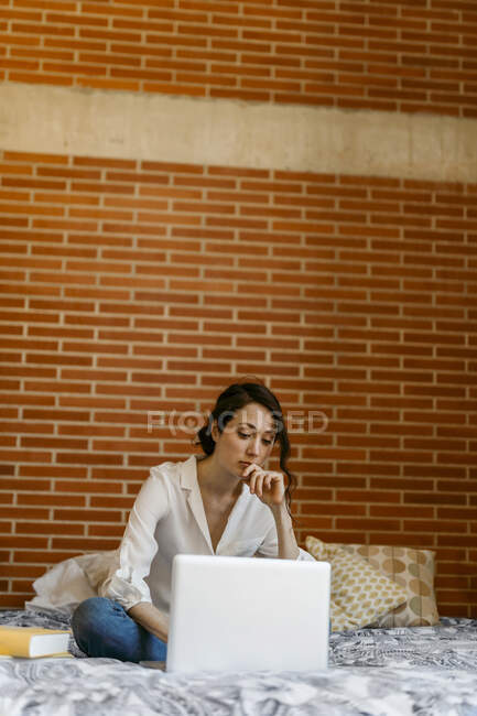 Задумчивая женщина с рукой на подбородке с помощью ноутбука, сидя перед кирпичной стеной — стоковое фото