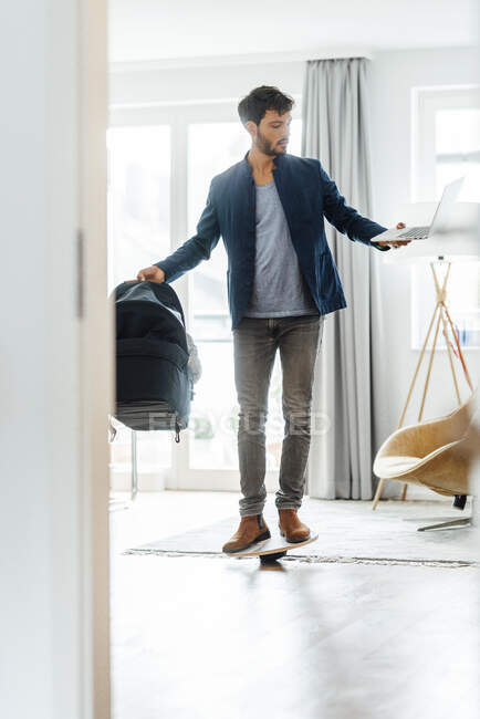 Male freelancer with baby carrier and laptop balancing on board in living room — Stock Photo